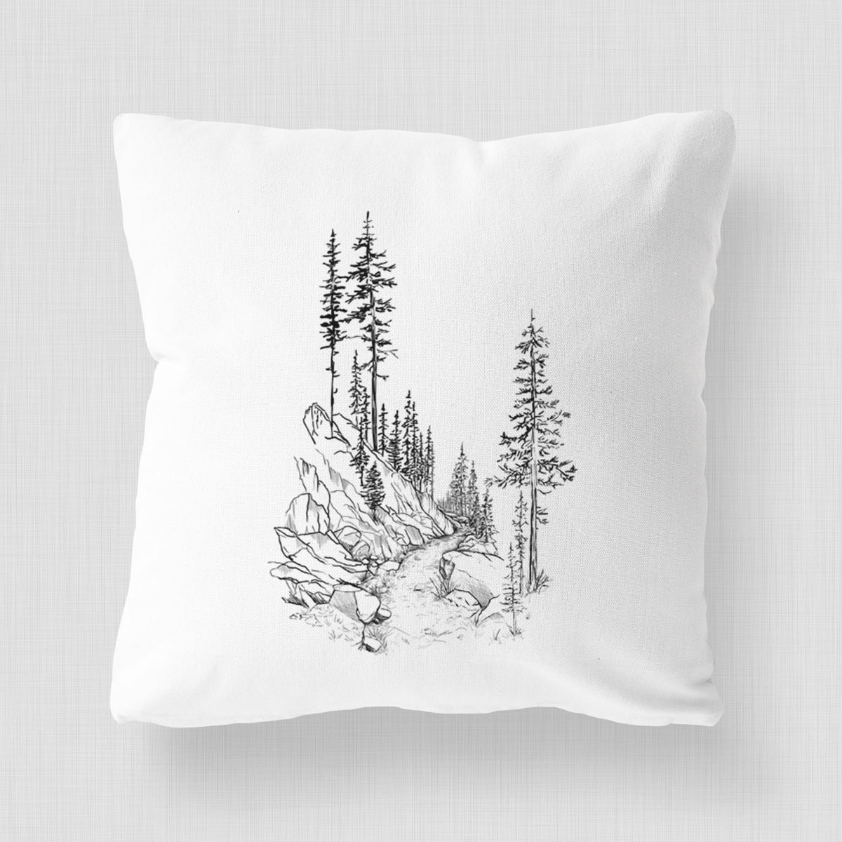 White pillow cover printed with a black ink illustration of a trail winding past tall pine trees and rocks into the woods.