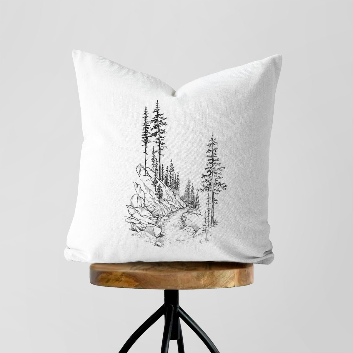 White pillow cover printed with a black ink illustration of a trail winding past tall pine trees and rocks into the woods, on a wooden stool.