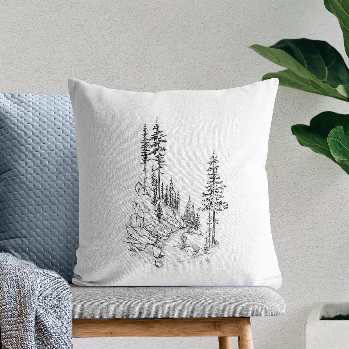 White pillow cover printed with a black ink illustration of a trail winding past tall pine trees and rocks into the woods, on a bench beside plants.