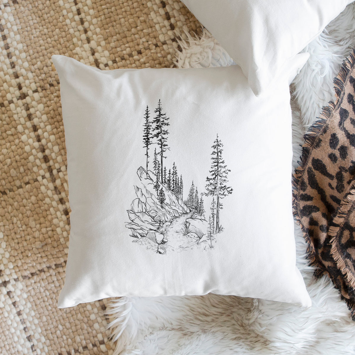 White pillow cover printed with a black ink illustration of a trail winding past tall pine trees and rocks into the woods, on a woven chair.