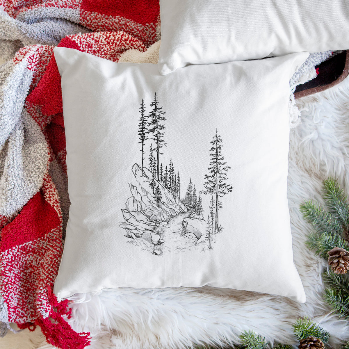 White pillow cover printed with a black ink illustration of a trail winding past tall pine trees and rocks into the woods, on a woven chair.