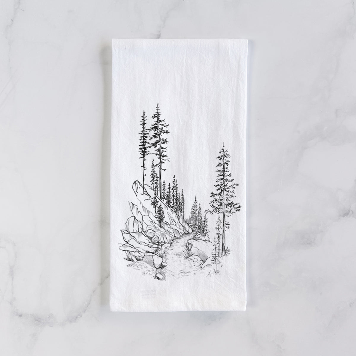 White cotton tea towel laid flat on marble, printed with a black ink illustration of a trail winding past tall pine trees and rocks into the woods.