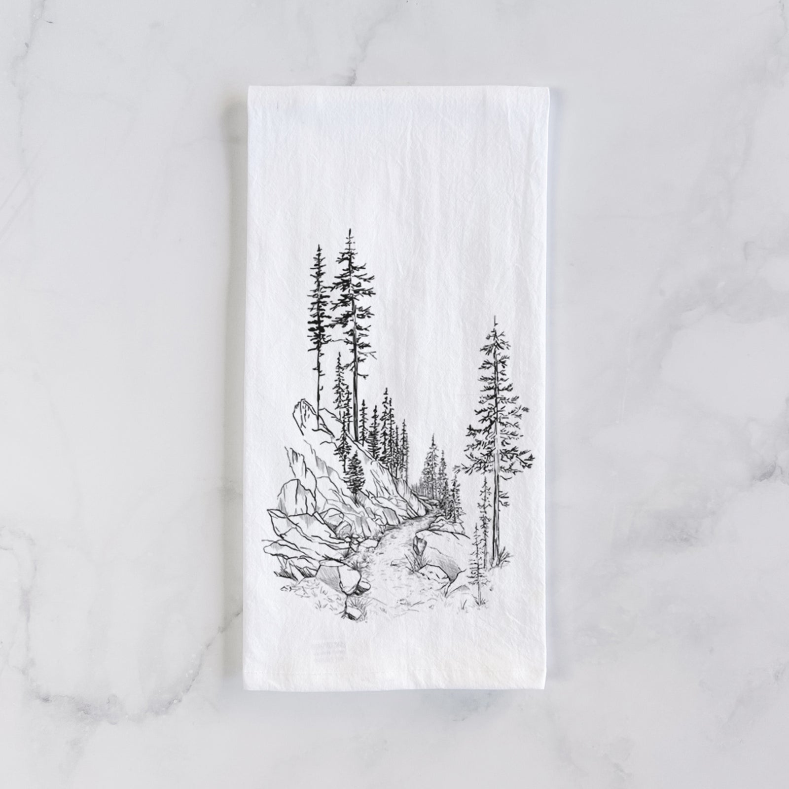 White cotton tea towel printed with a black ink illustration of a trail winding past tall pine trees and rocks into the woods, on a marble counter beside a round wooden cutting board and greenery.