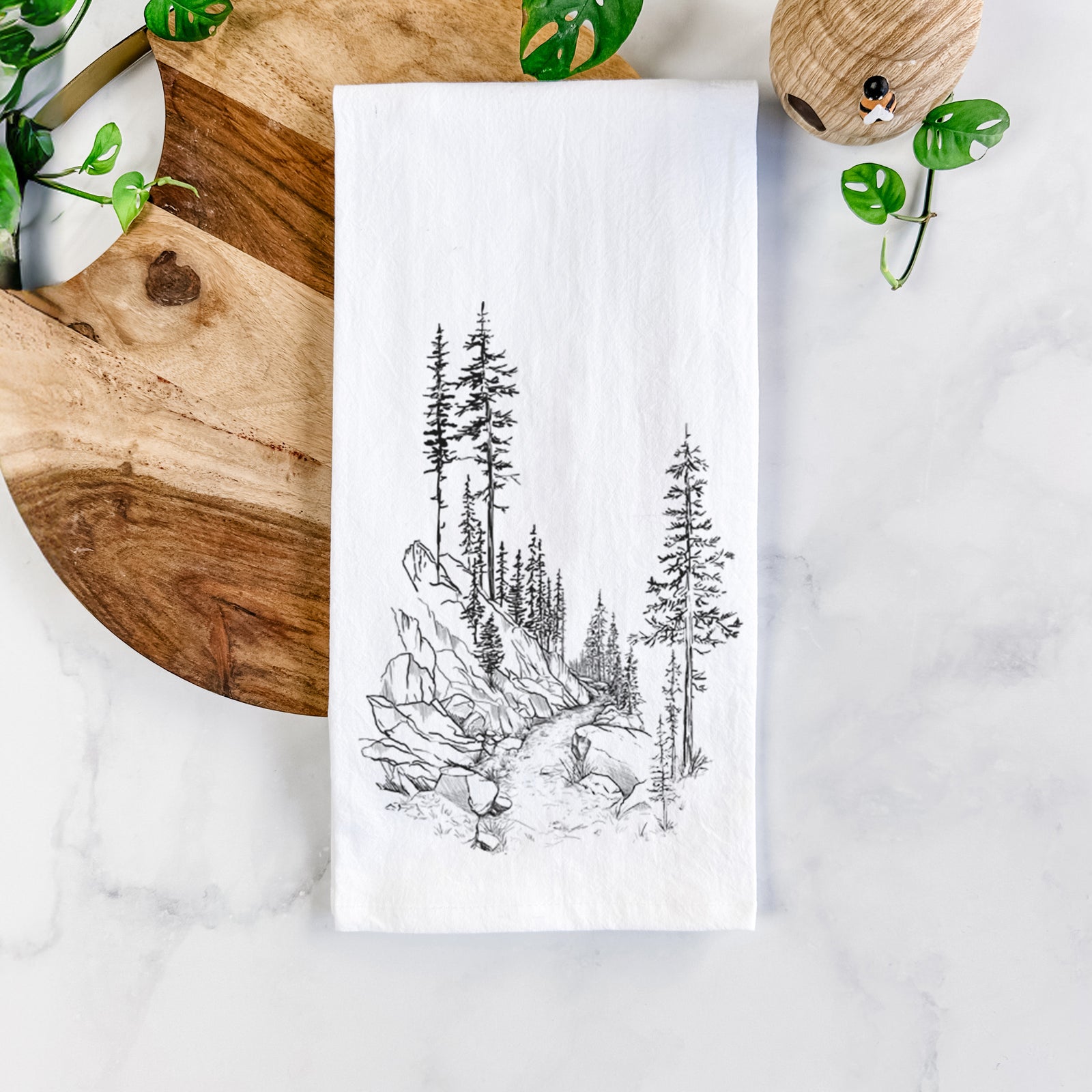 White cotton tea towel printed with a black ink illustration of a trail winding past tall pine trees and rocks into the woods, on a marble counter beside a round wooden cutting board and greenery.