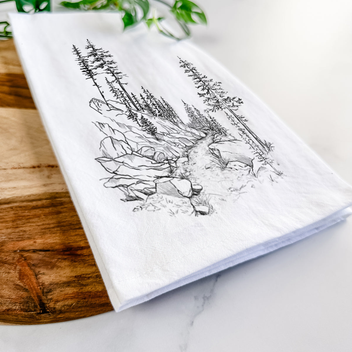 Close-up of a folded white tea towel on a wooden board, showing a black ink illustration of a trail winding past tall pine trees and rocks into the woods.