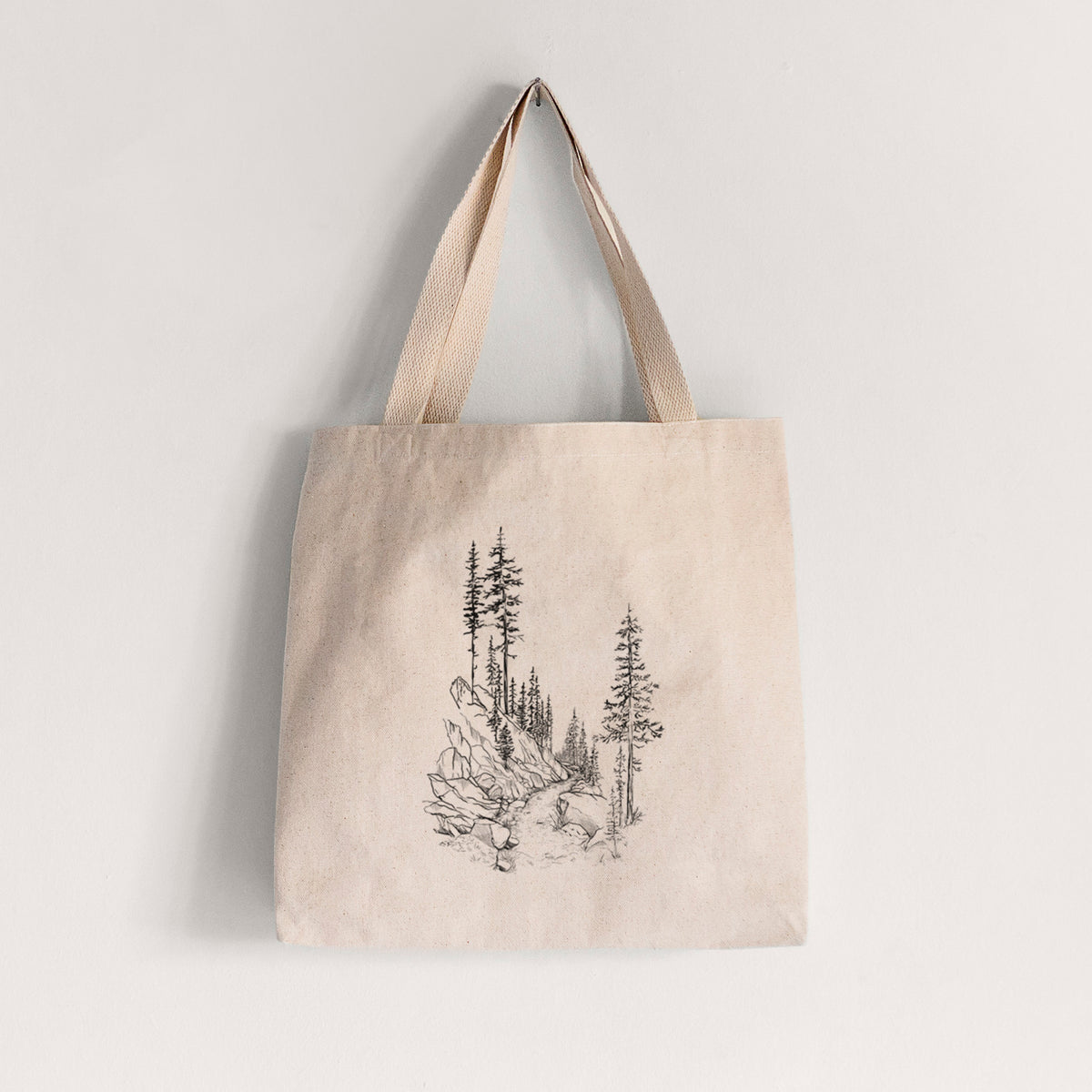 Natural canvas tote bag printed with a black ink illustration of a trail winding past tall pine trees and rocks into the woods.
