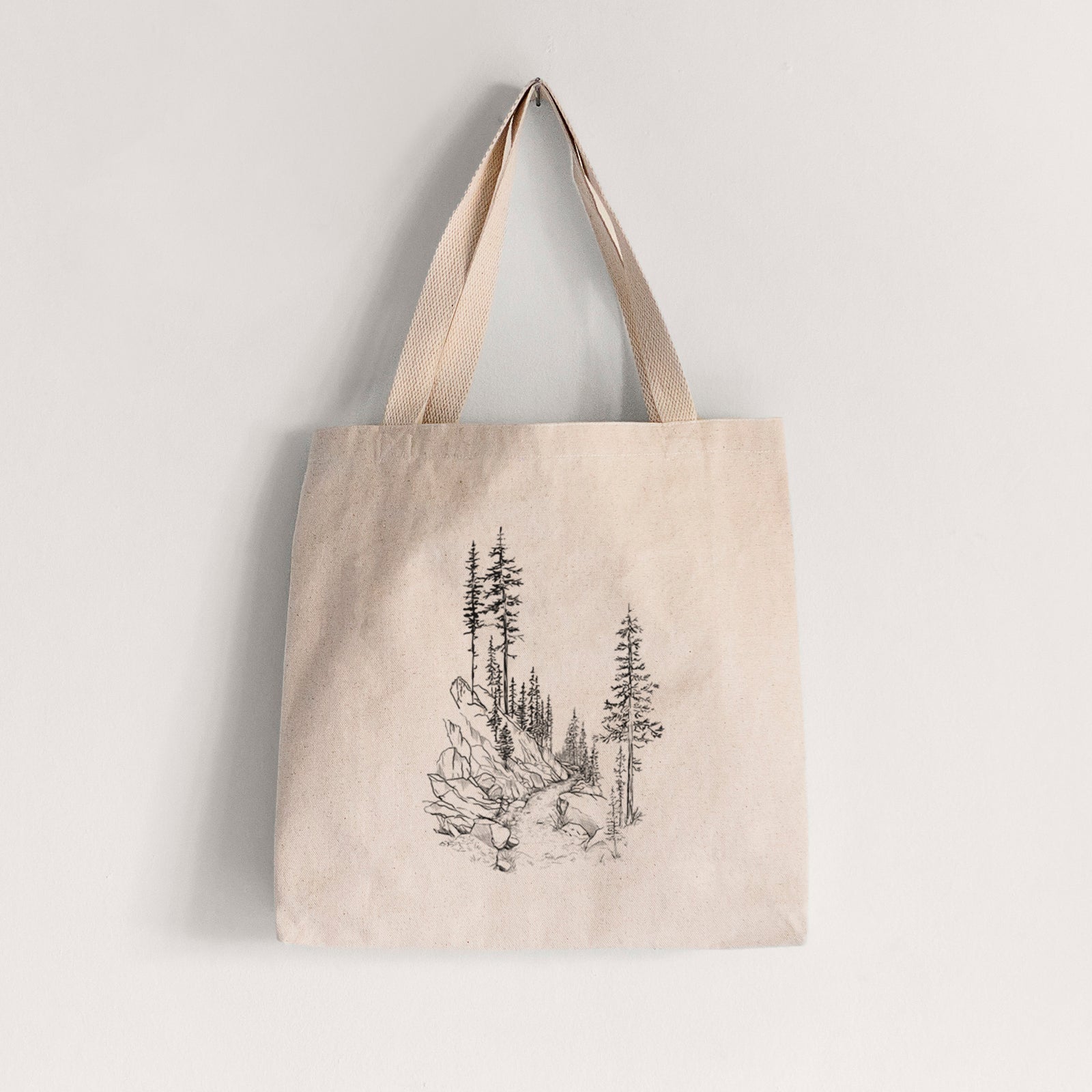 Natural canvas tote bag printed with a black ink illustration of a trail winding past tall pine trees and rocks into the woods.