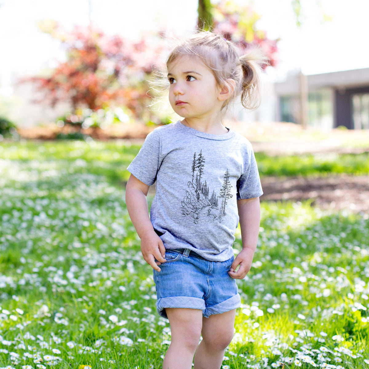 Toddler girl walking outdoors wearing a t-shirt printed with a black ink illustration of a trail winding past tall pine trees and rocks into the woods.