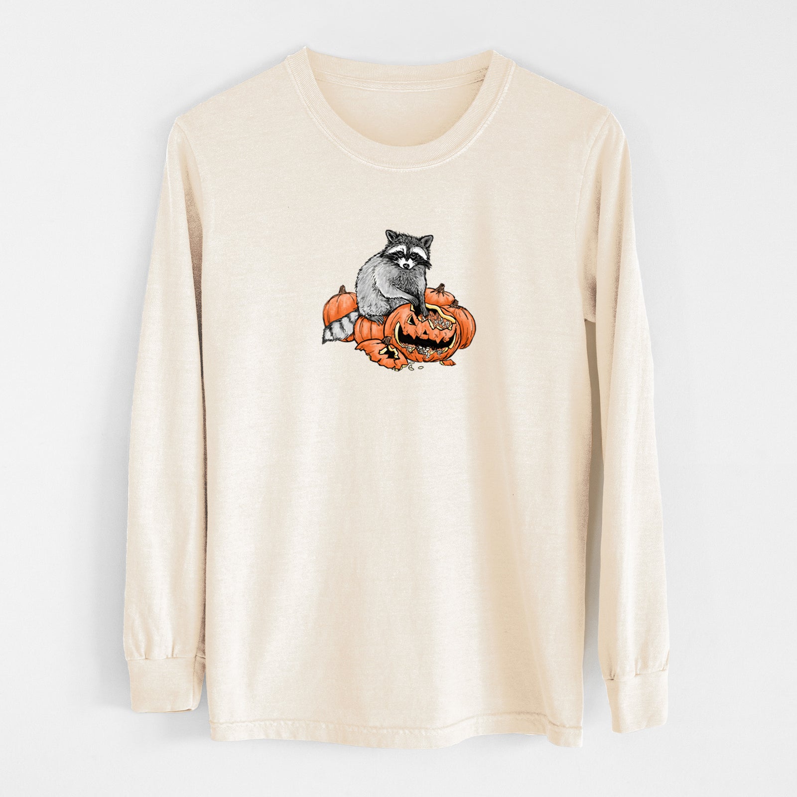 Ivory Comfort Colors long sleeve shirt laid flat, printed with a black ink raccoon peeking over carved orange jack-o'-lanterns.