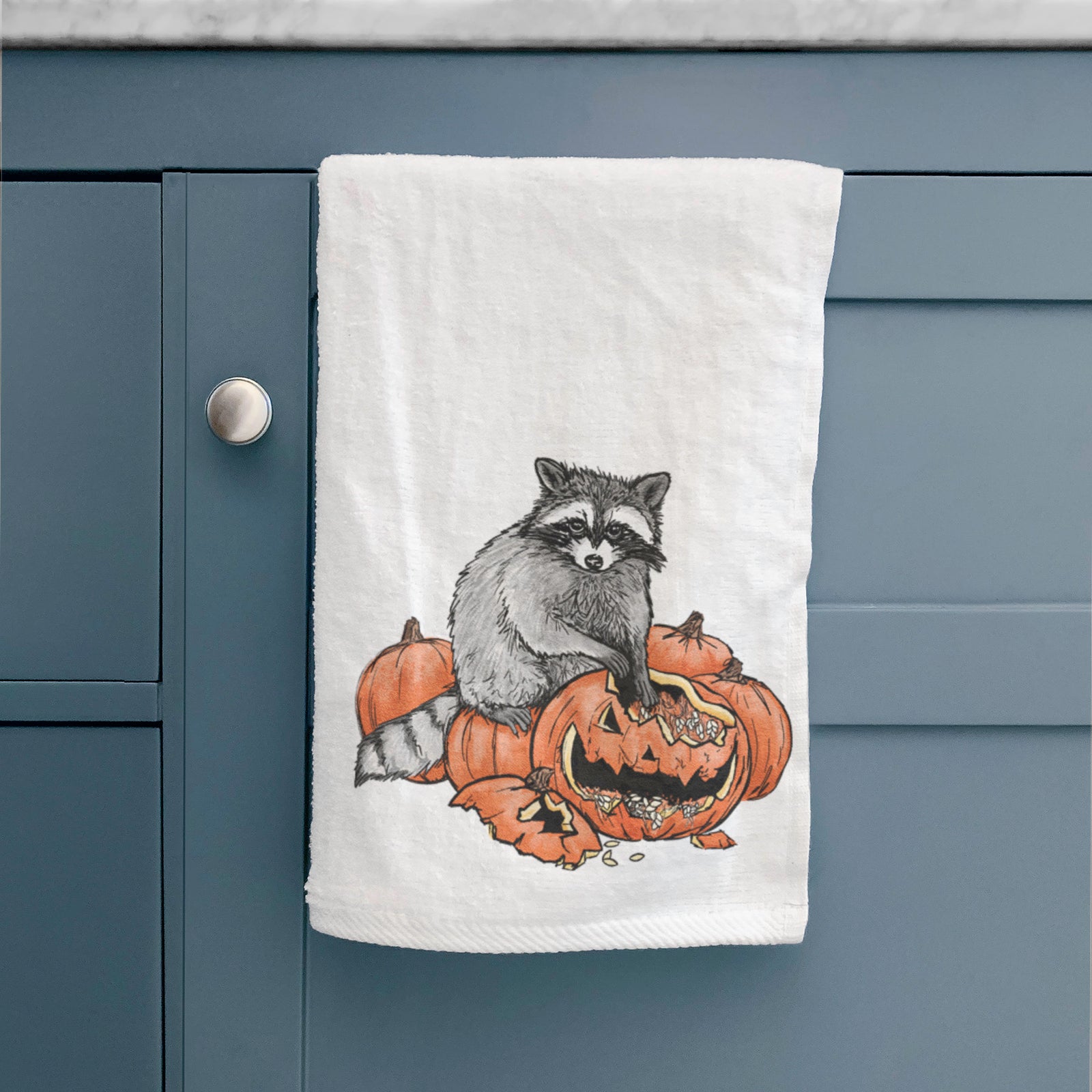White hand towel printed with a black ink raccoon peeking over carved orange jack-o'-lanterns.