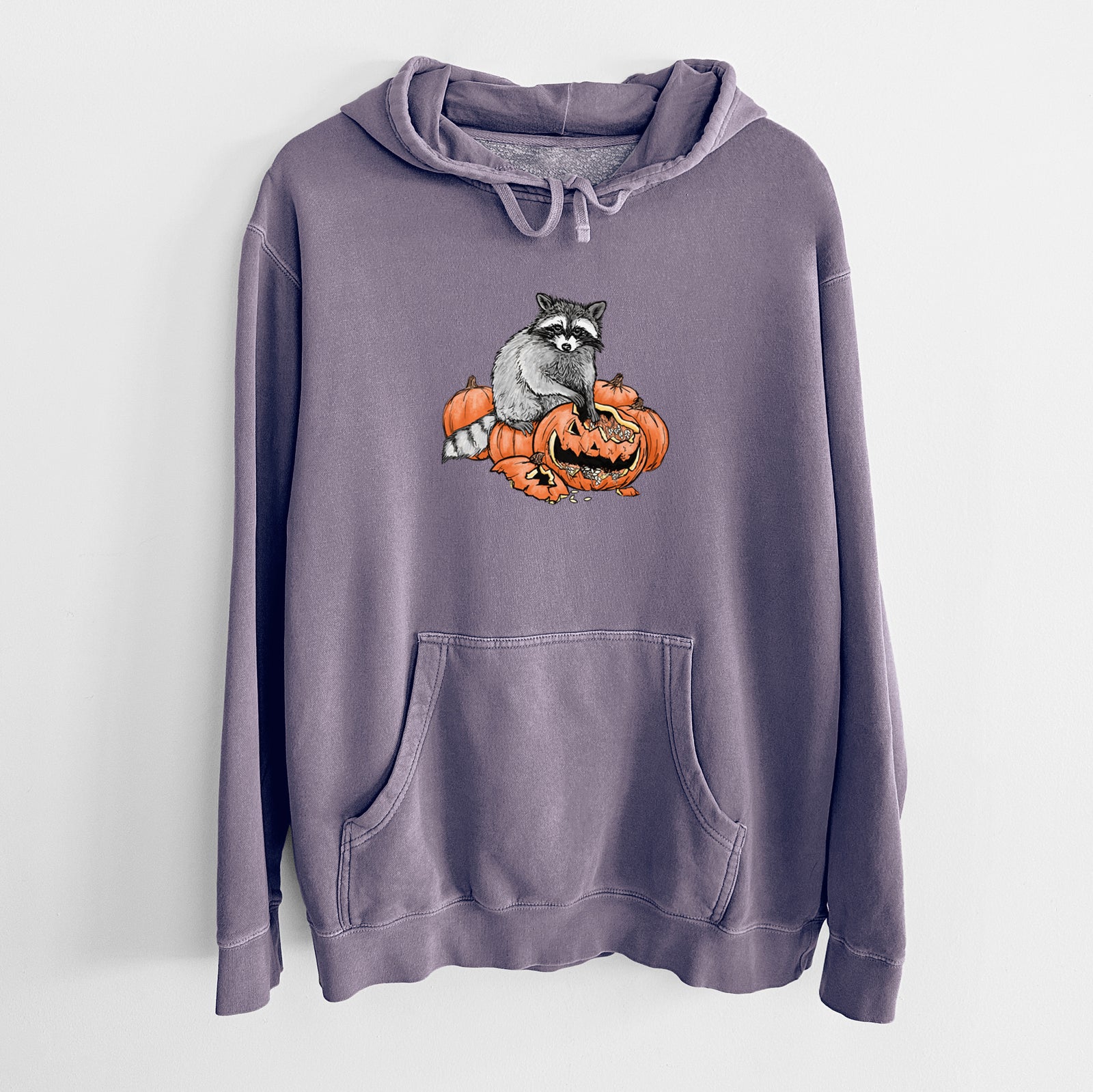 Plum purple pigment dyed hoodie laid flat, printed with a black ink raccoon peeking over carved orange jack-o'-lanterns.