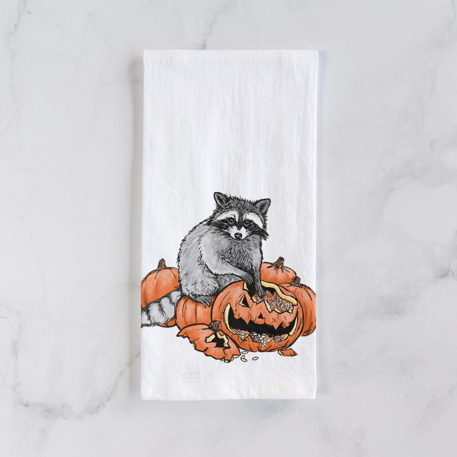 White cotton tea towel printed with a black ink raccoon peeking over carved orange jack-o'-lanterns, on a marble counter beside a round wooden cutting board and greenery.