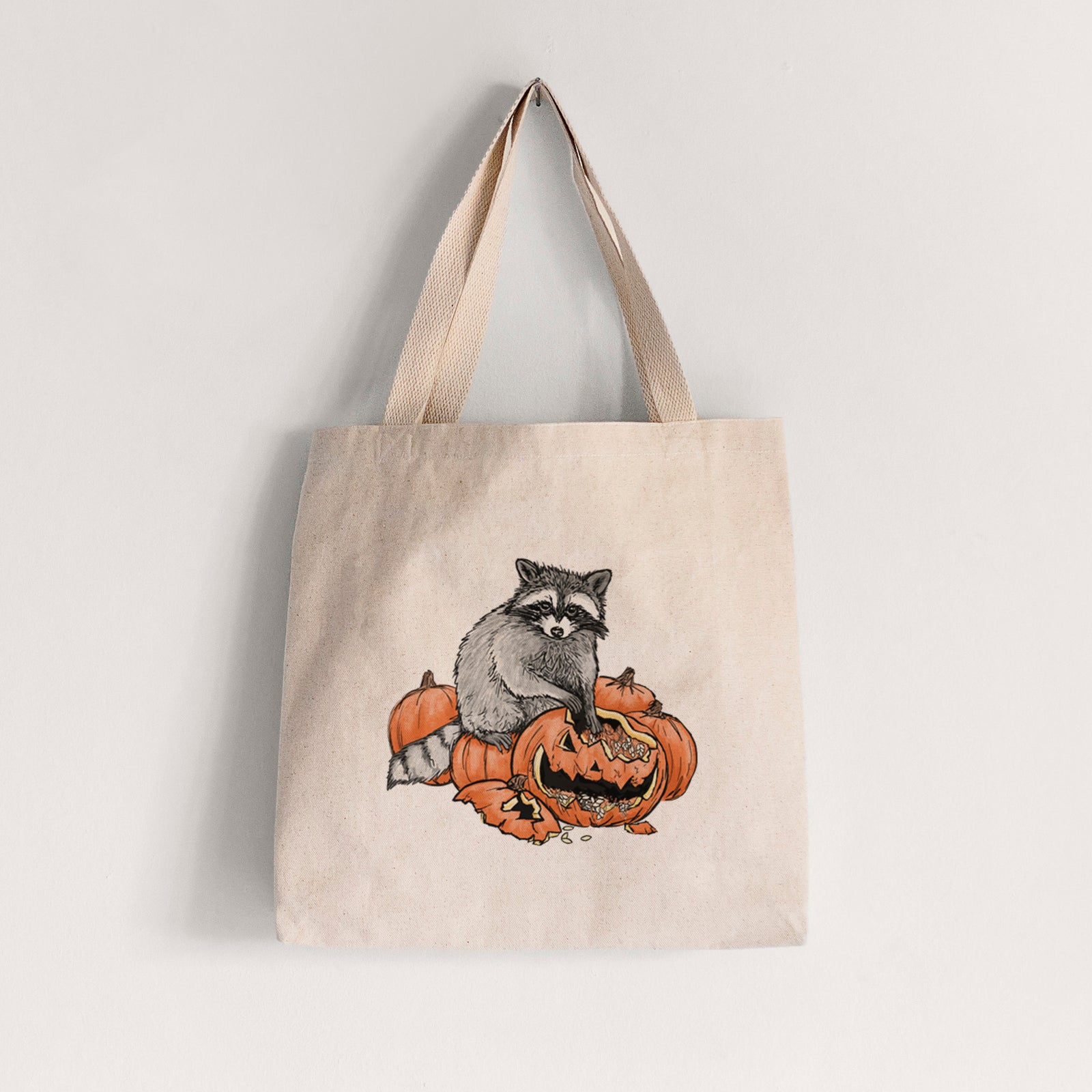 Natural canvas tote bag printed with a black ink raccoon peeking over carved orange jack-o'-lanterns.
