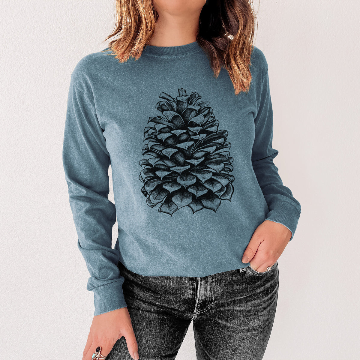 Steel blue Comfort Colors long sleeve shirt printed with a detailed black ink illustration of a large Jeffrey pine cone, shown in a lifestyle photo.