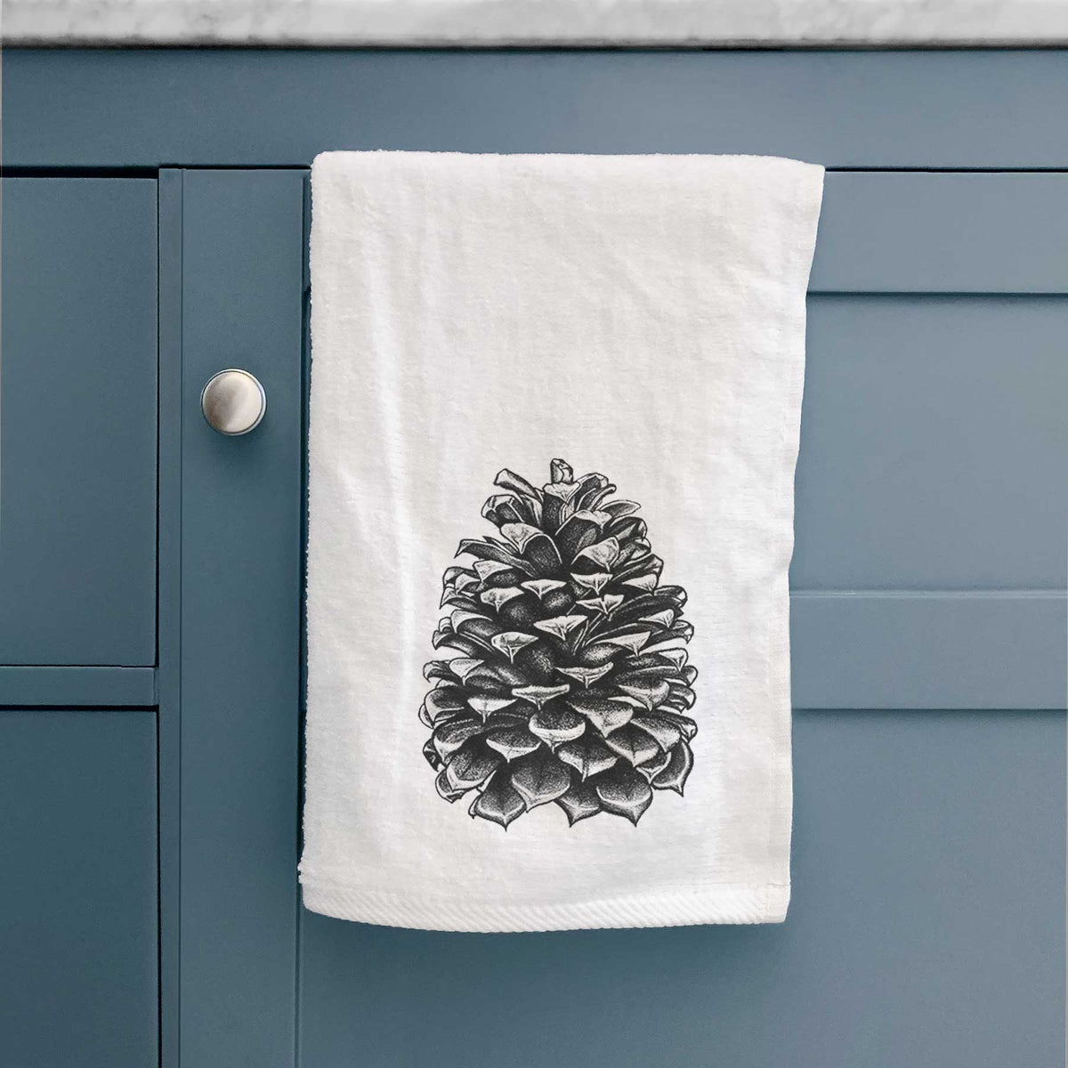 White hand towel printed with a detailed black ink illustration of a large Jeffrey pine cone, hanging on a cabinet door.