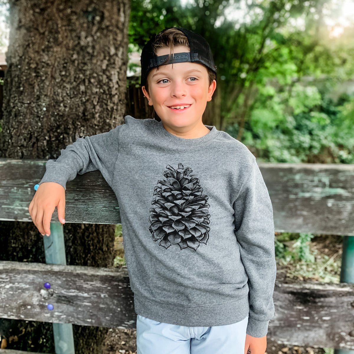 Boy wearing a youth crewneck sweatshirt printed with a detailed black ink illustration of a large Jeffrey pine cone, outdoors.