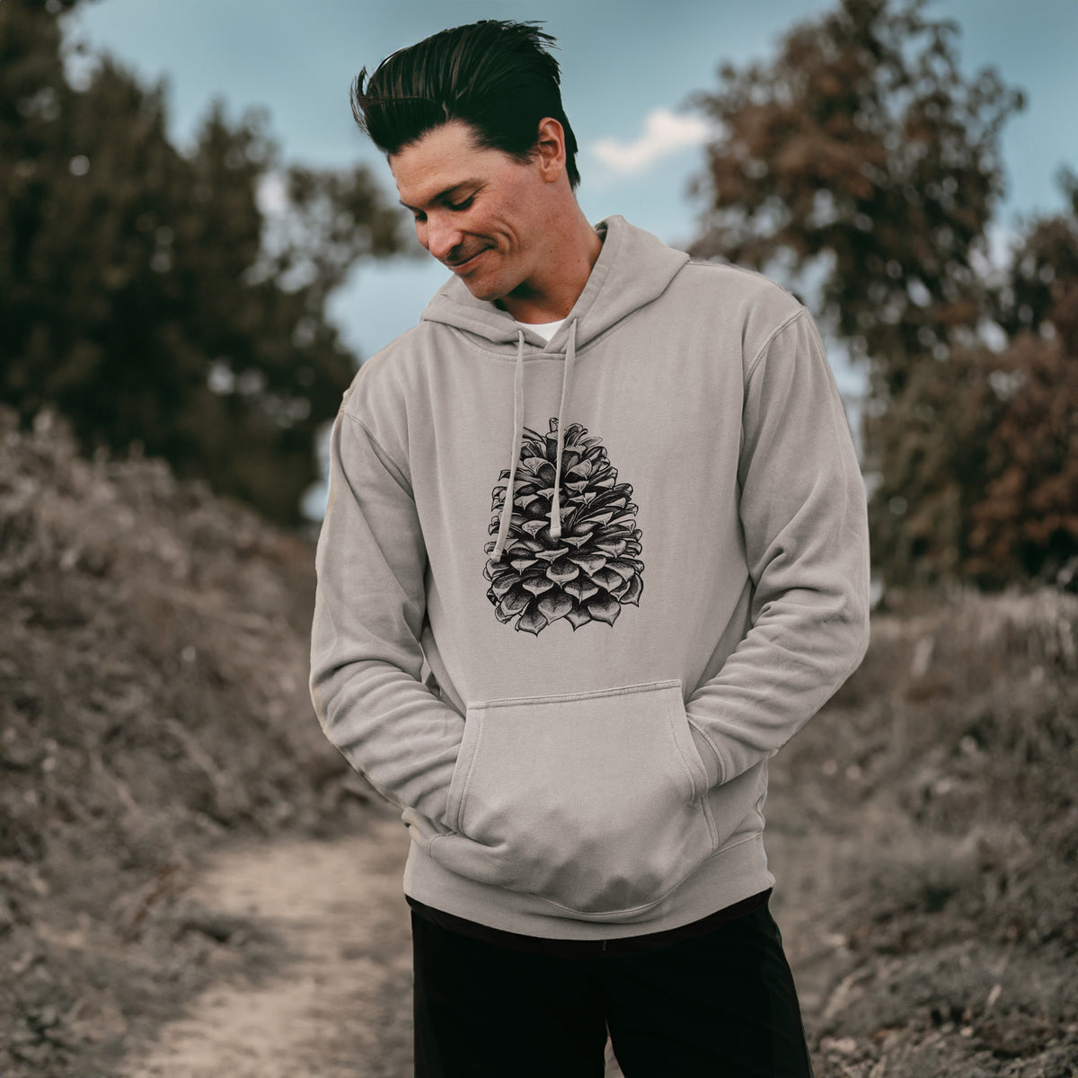 Man wearing a cement grey pigment dyed hoodie printed with a detailed black ink illustration of a large Jeffrey pine cone.