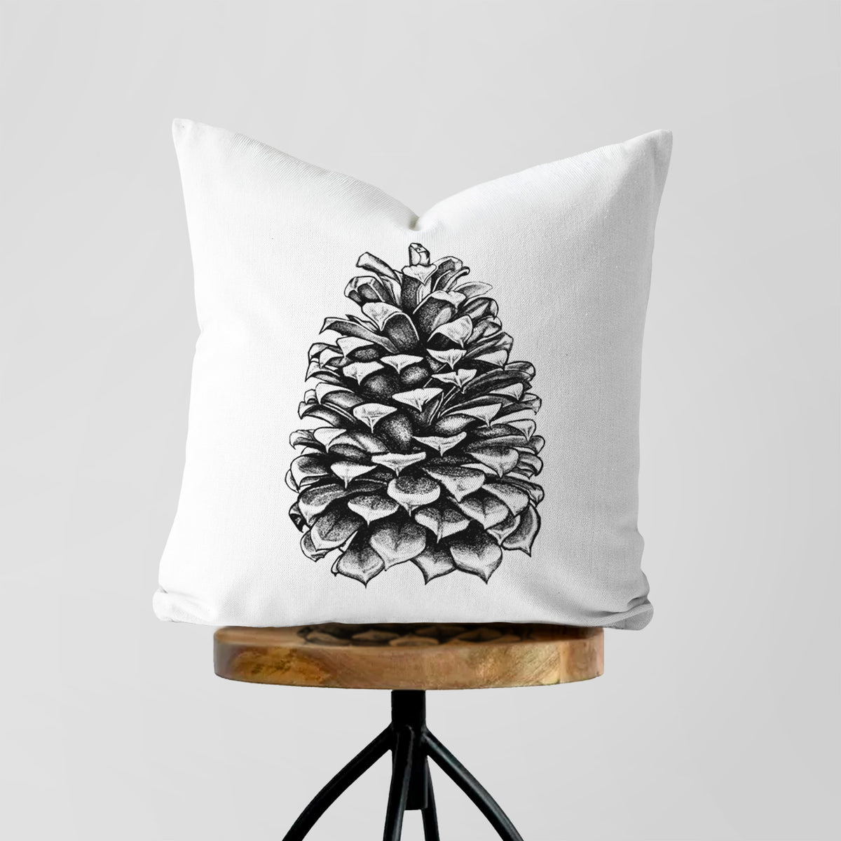 White pillow cover printed with a detailed black ink illustration of a large Jeffrey pine cone, on a wooden stool.