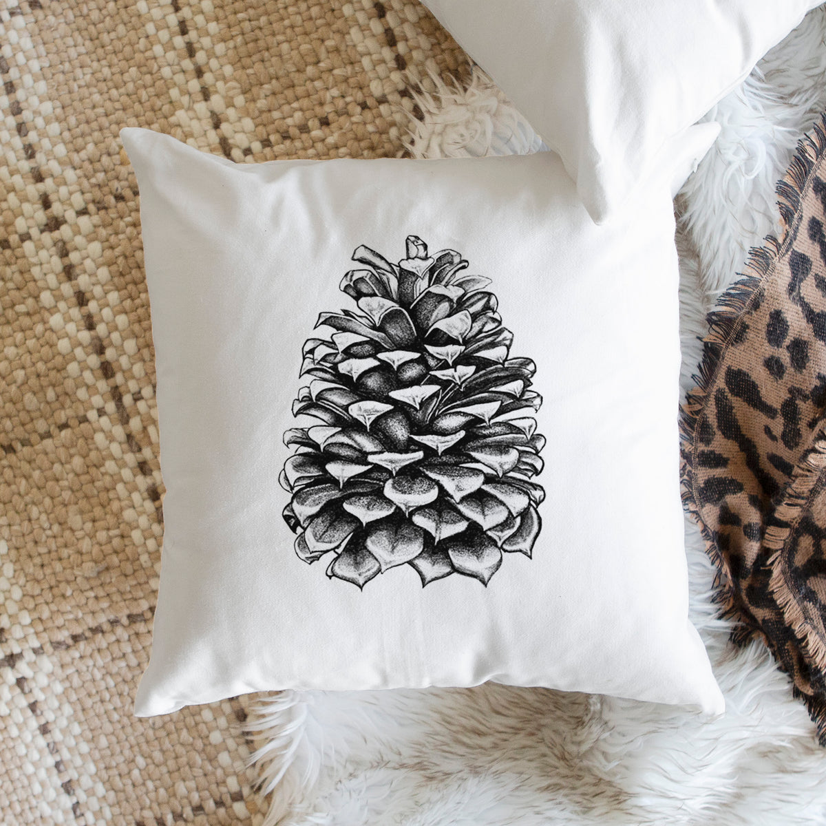 White pillow cover printed with a detailed black ink illustration of a large Jeffrey pine cone, on a woven chair.