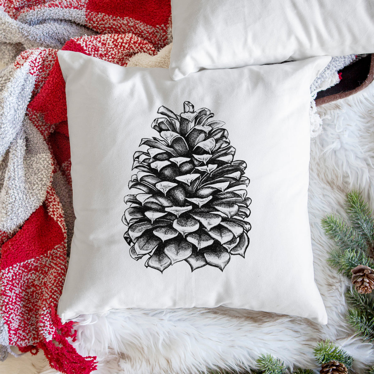White pillow cover printed with a detailed black ink illustration of a large Jeffrey pine cone, on a woven chair.
