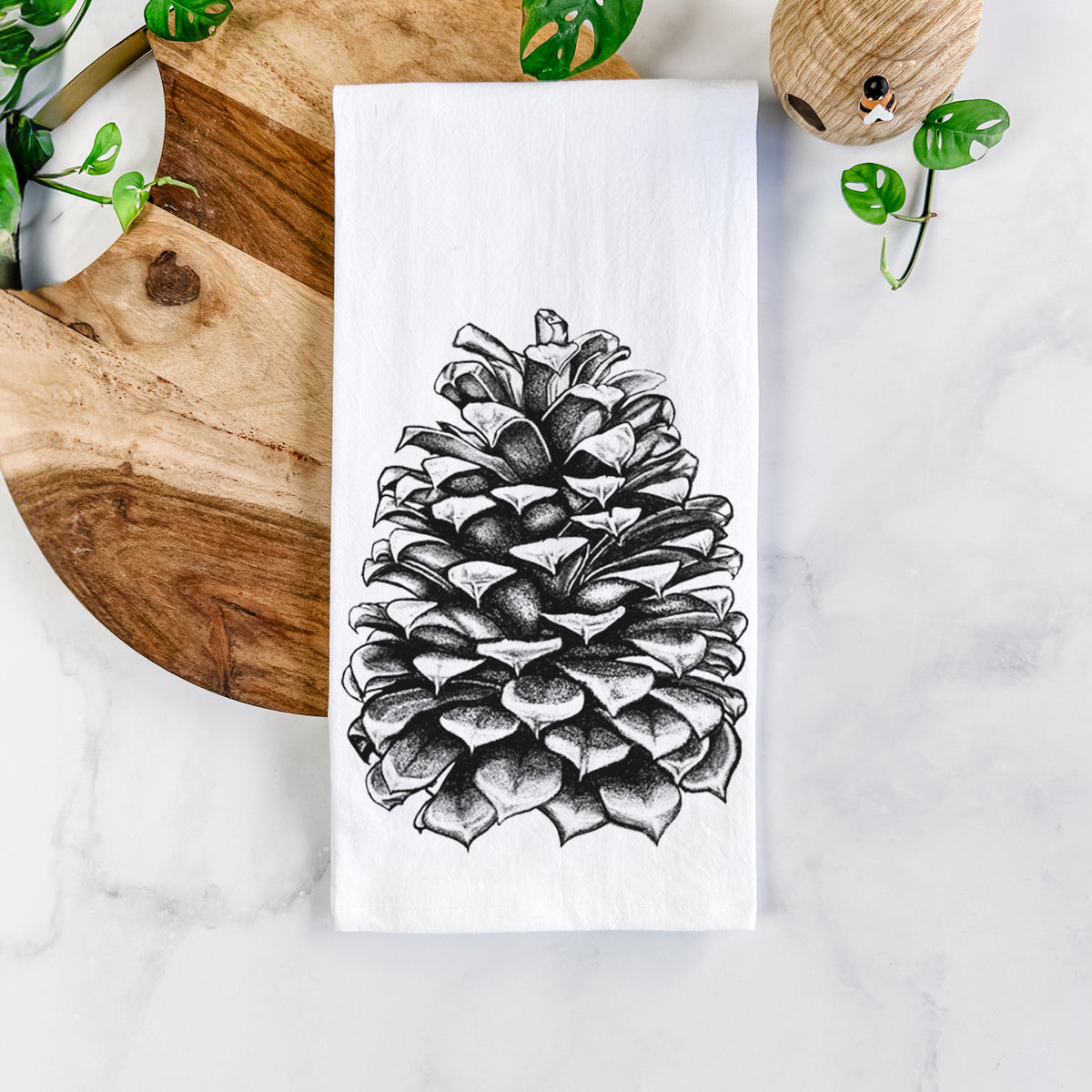 White cotton tea towel printed with a detailed black ink illustration of a large Jeffrey pine cone, on a marble counter beside a round wooden cutting board and greenery.