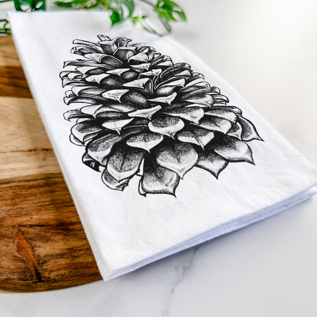Close-up of a folded white tea towel on a wooden board, showing a detailed black ink illustration of a large Jeffrey pine cone.