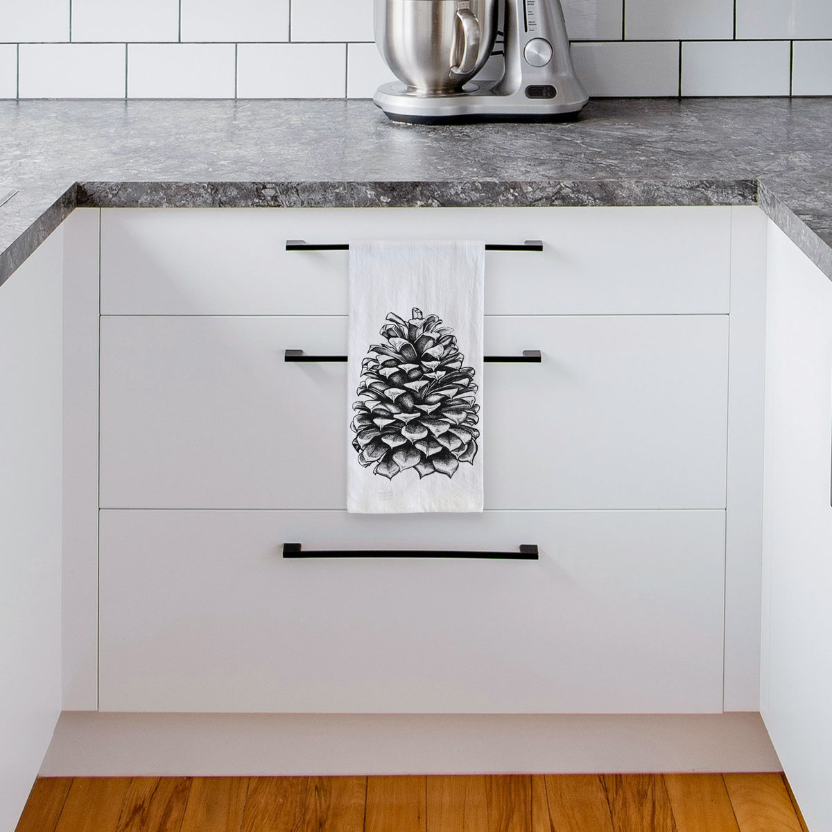 White tea towel printed with a detailed black ink illustration of a large Jeffrey pine cone, hanging from a drawer handle in a bright white kitchen.