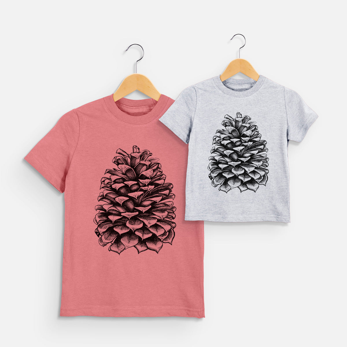 Two youth t-shirts in different colors on hangers, printed with a detailed black ink illustration of a large Jeffrey pine cone.