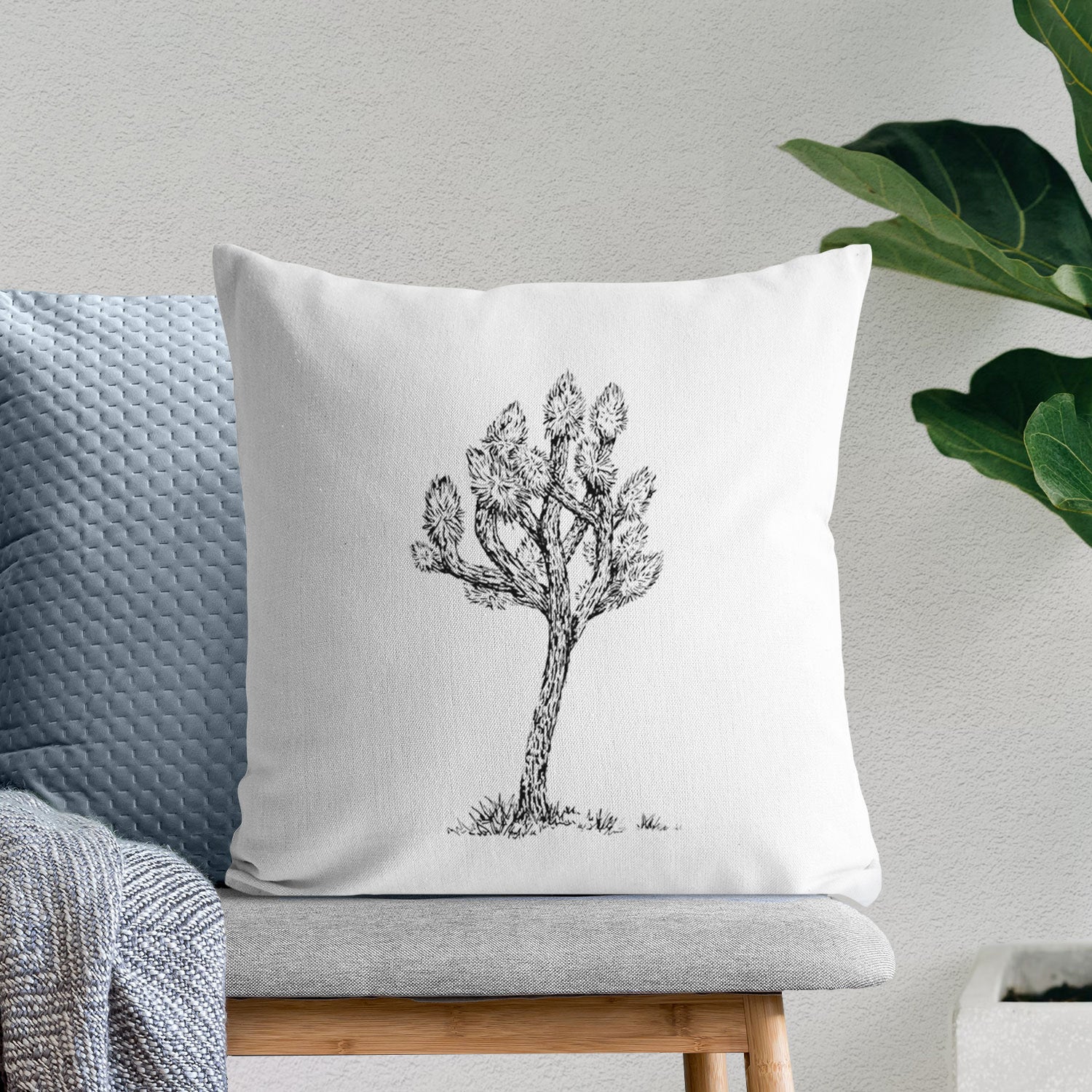 White pillow cover printed with a detailed black ink illustration of a Joshua tree, on a bench beside plants.