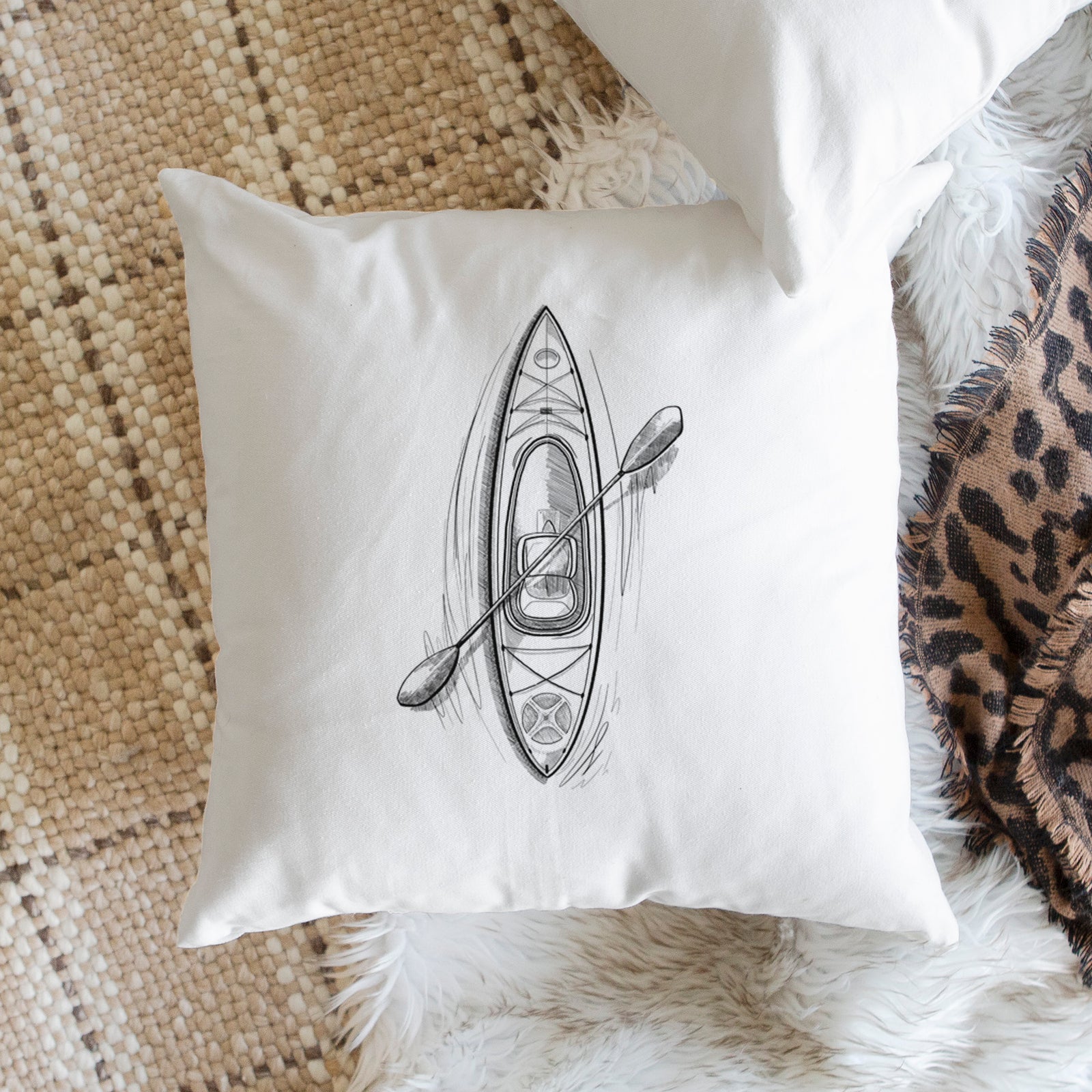 White pillow cover printed with a black ink illustration of a kayak with a paddle, seen from above, on a bench beside plants.