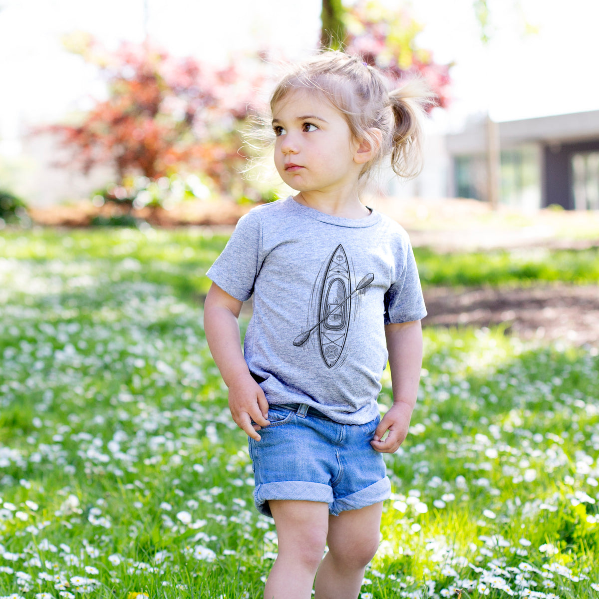 Toddler girl walking outdoors wearing a t-shirt printed with a black ink illustration of a kayak with a paddle, seen from above.