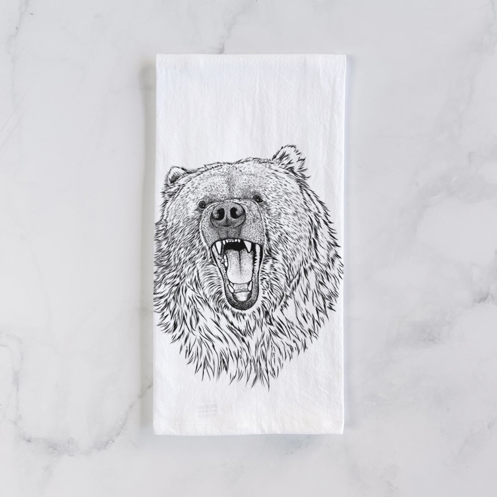 White cotton tea towel printed with a detailed black ink portrait of a roaring Kodiak bear facing forward, on a marble counter beside a round wooden cutting board and greenery.