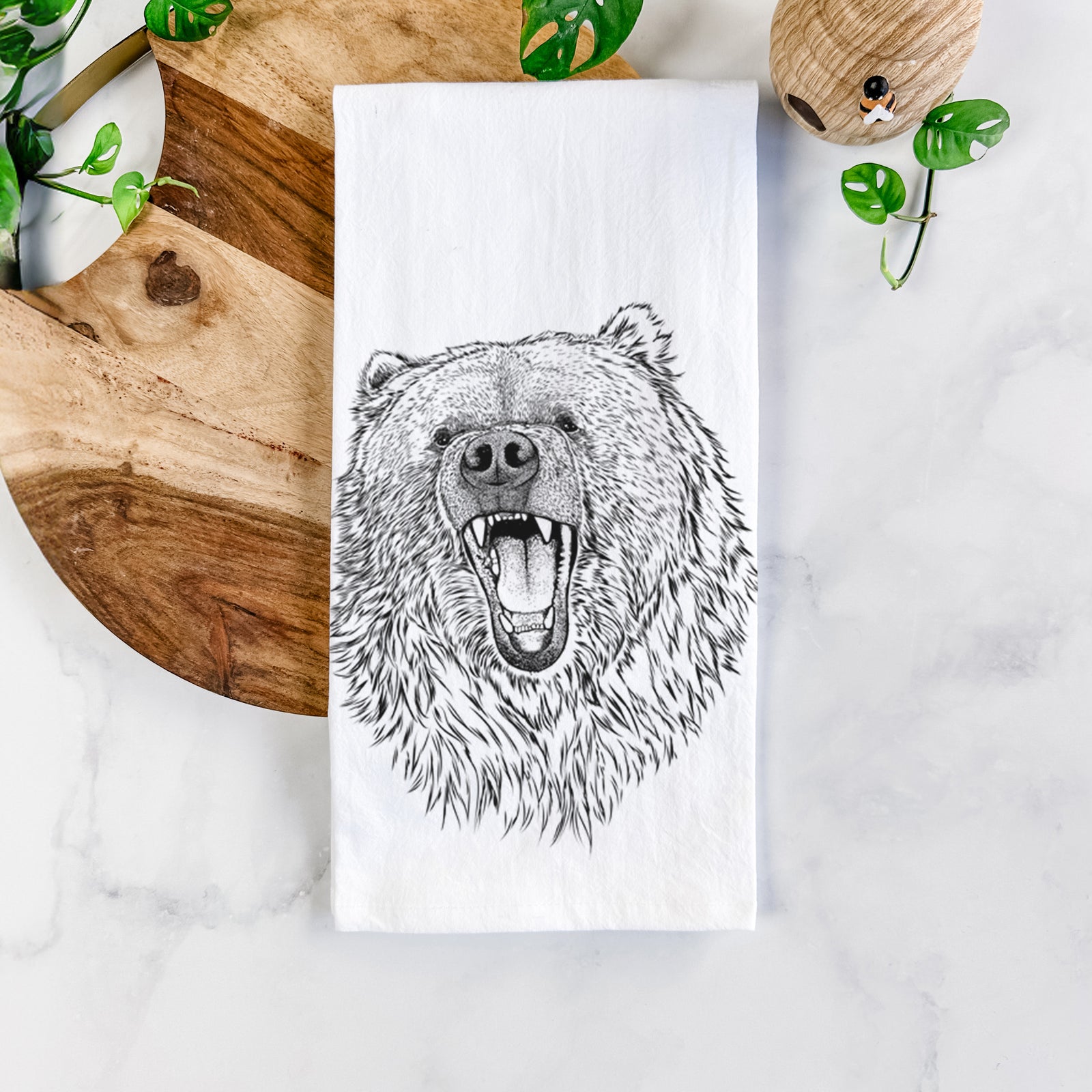 White cotton tea towel printed with a detailed black ink portrait of a roaring Kodiak bear facing forward, on a marble counter beside a round wooden cutting board and greenery.