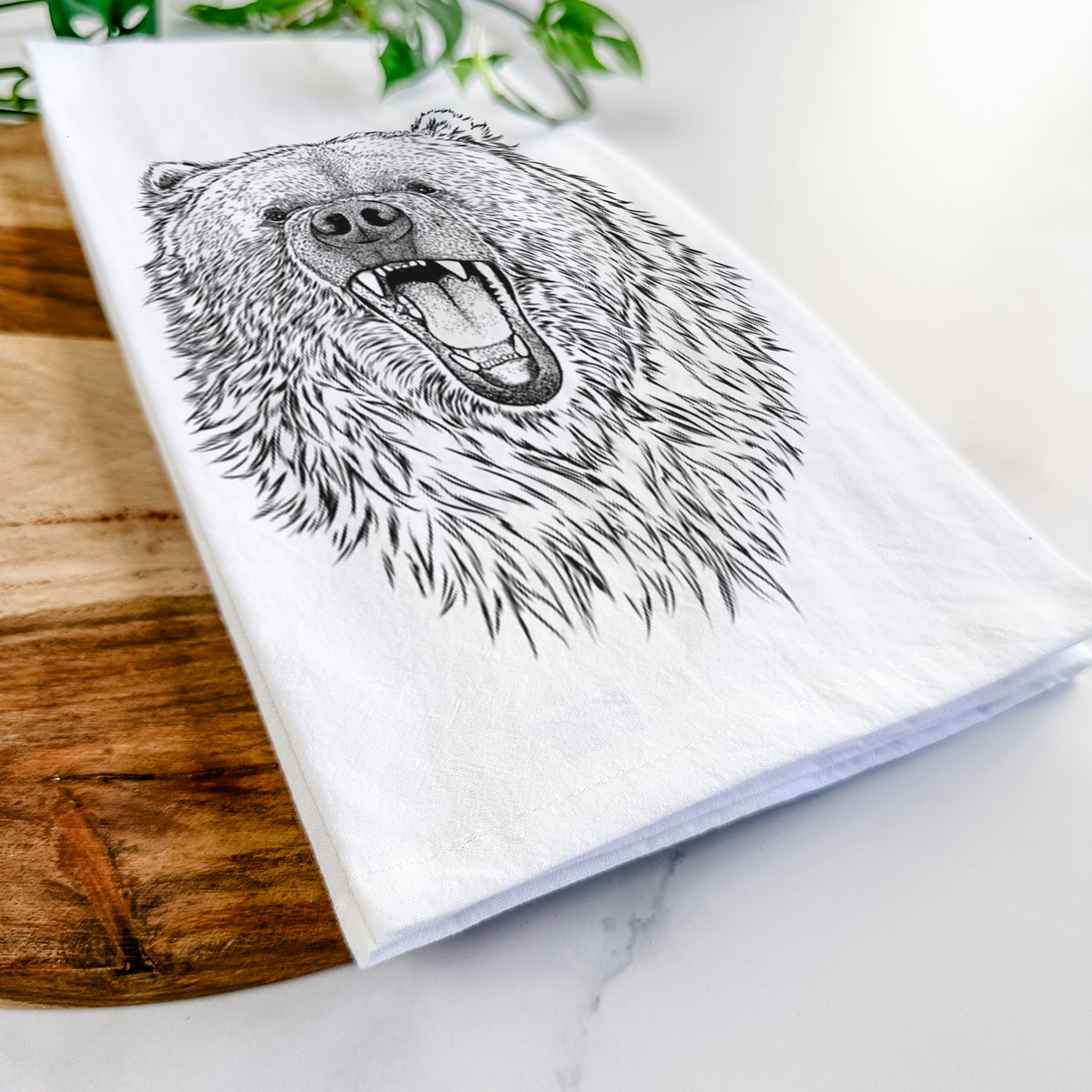 Close-up of a folded white tea towel on a wooden board, showing a detailed black ink portrait of a roaring Kodiak bear facing forward.
