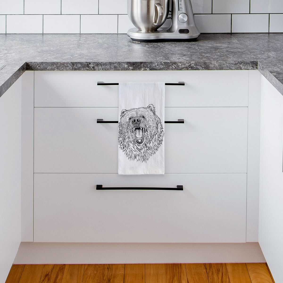 White tea towel printed with a detailed black ink portrait of a roaring Kodiak bear facing forward, hanging from a drawer handle in a bright white kitchen.