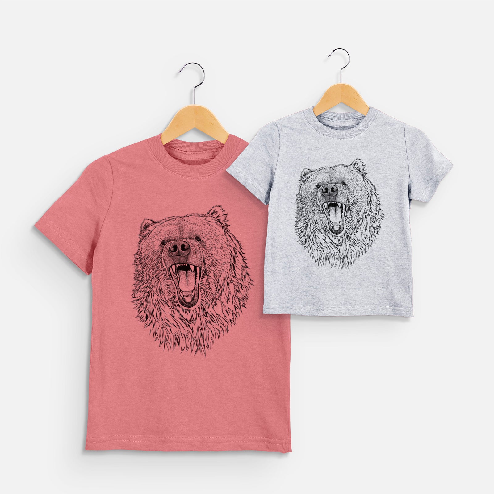 Two youth t-shirts in different colors on hangers, printed with a detailed black ink portrait of a roaring Kodiak bear facing forward.