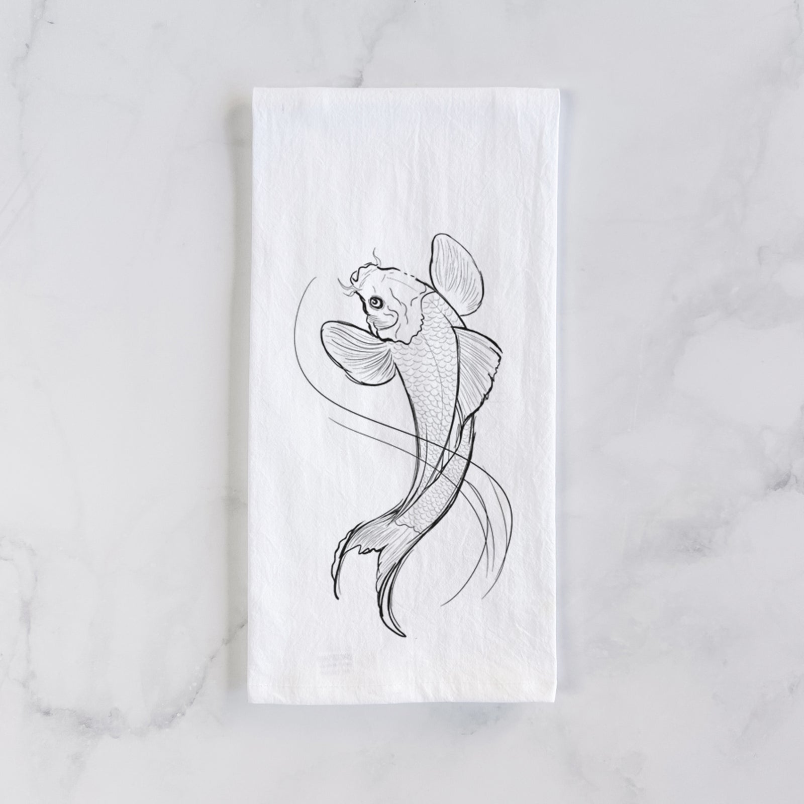 White cotton tea towel printed with a black ink line drawing of a koi fish swimming with flowing fins, on a marble counter beside a round wooden cutting board and greenery.