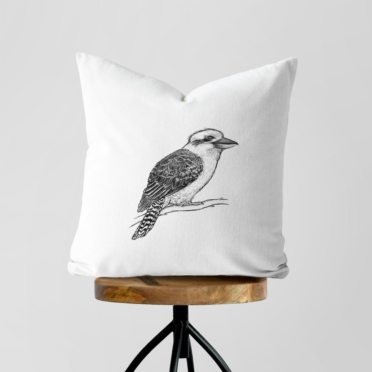 White pillow cover printed with a detailed black ink sketch of a kookaburra perched on a branch, on a wooden stool.