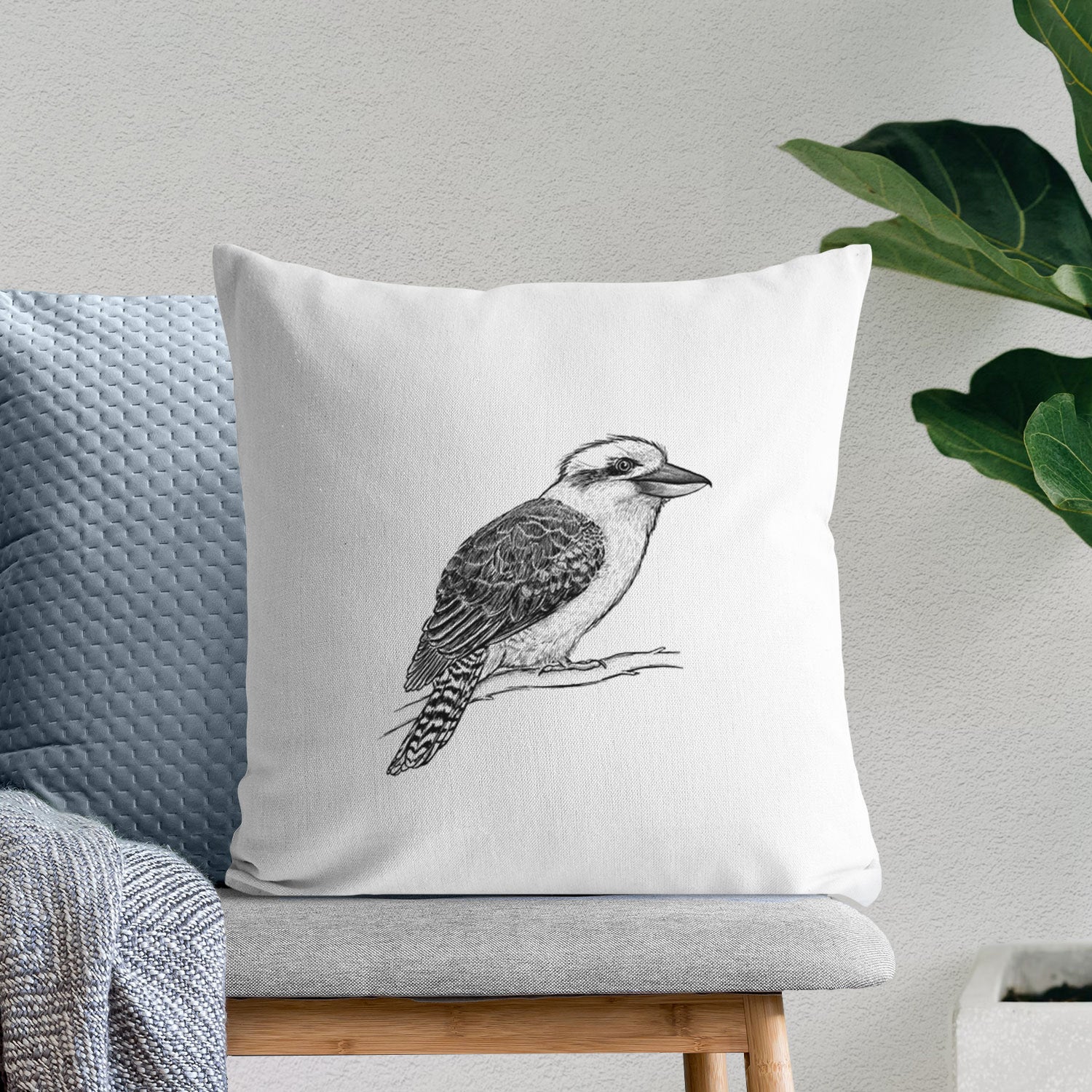 White pillow cover printed with a detailed black ink sketch of a kookaburra perched on a branch, on a bench beside plants.