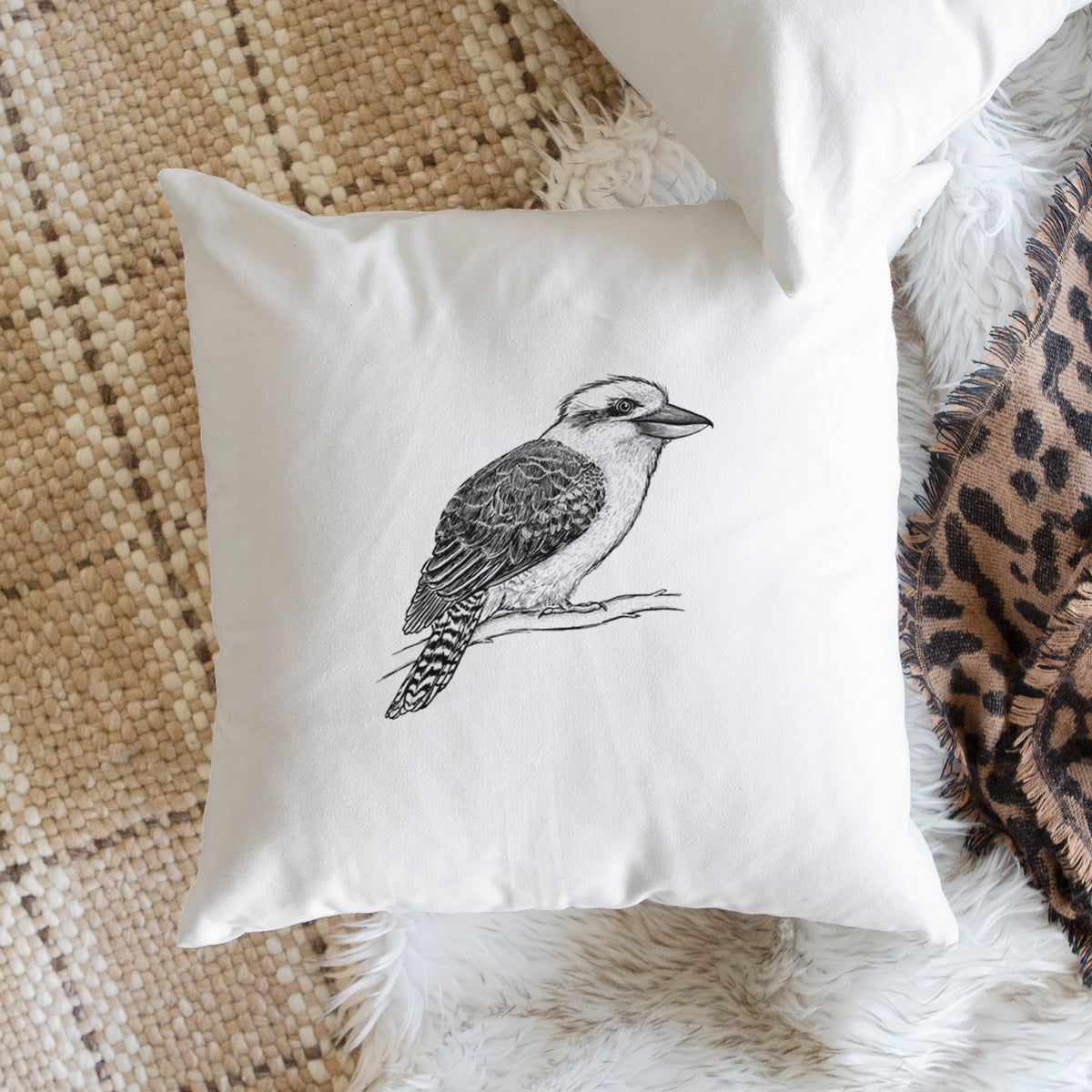 White pillow cover printed with a detailed black ink sketch of a kookaburra perched on a branch, on a woven chair.