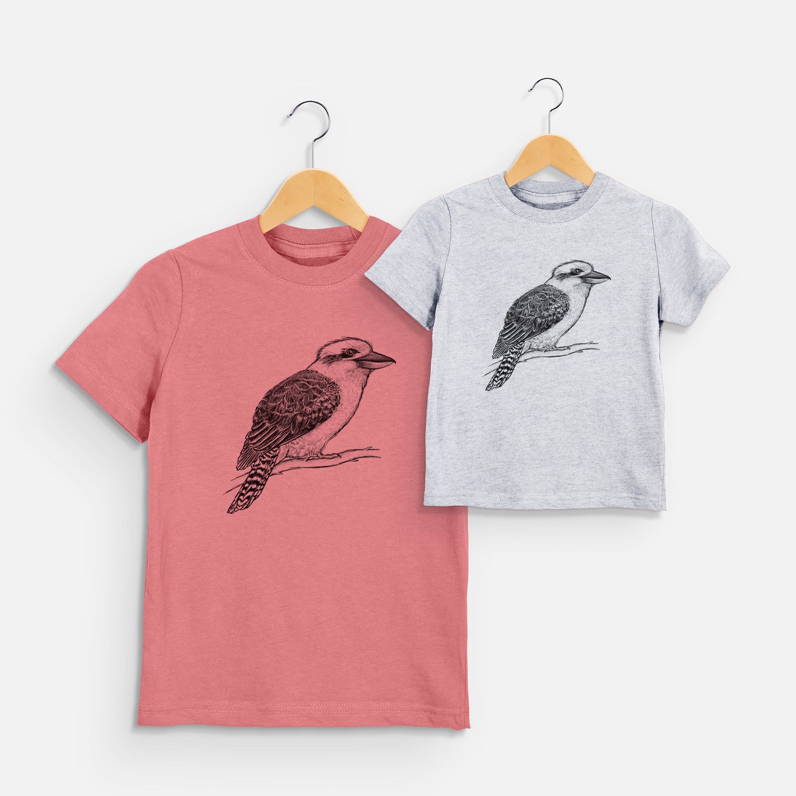 Two youth t-shirts in different colors on hangers, printed with a detailed black ink sketch of a kookaburra perched on a branch.