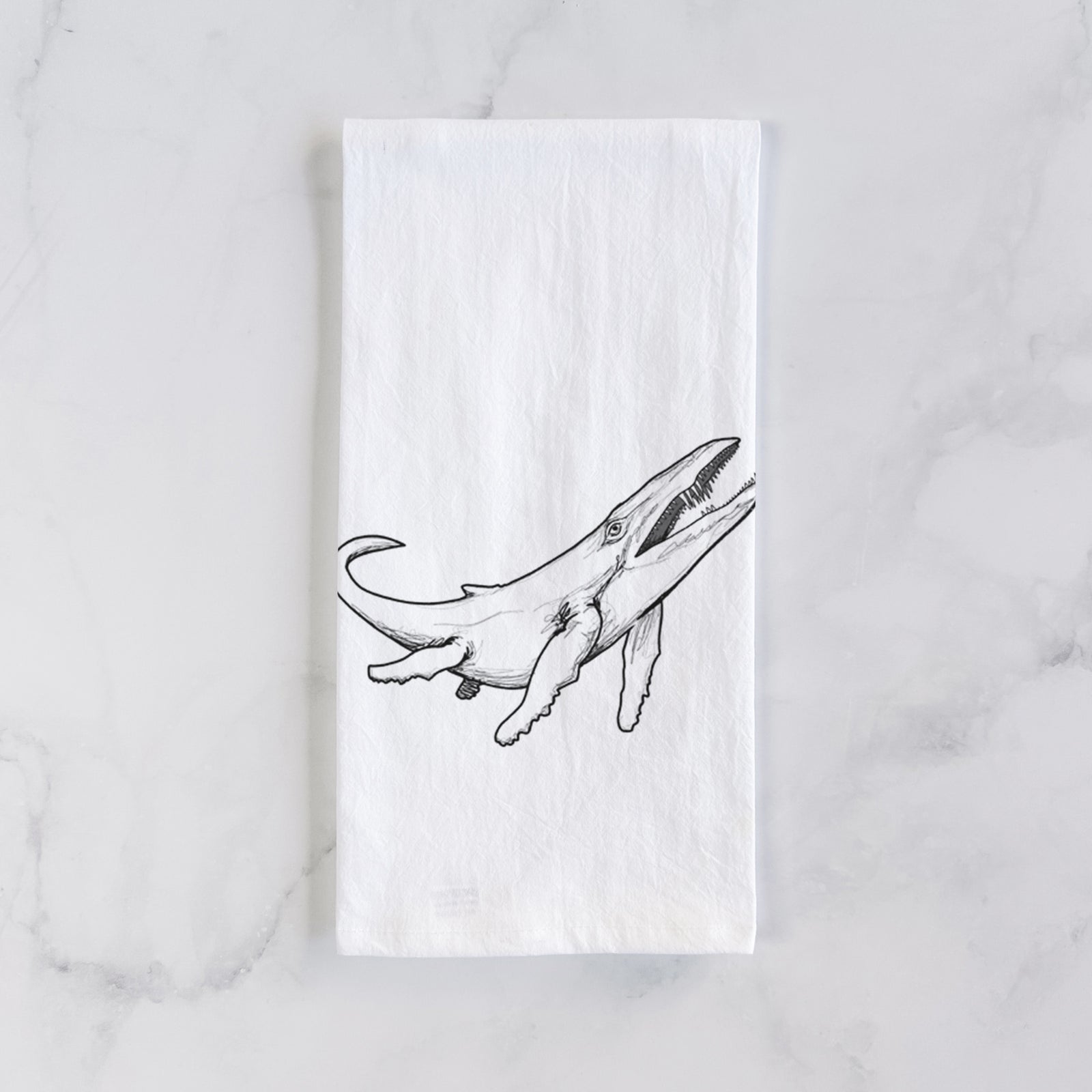 White cotton tea towel printed with a black ink illustration of a Kronosaurus marine reptile swimming, on a marble counter beside a round wooden cutting board and greenery.