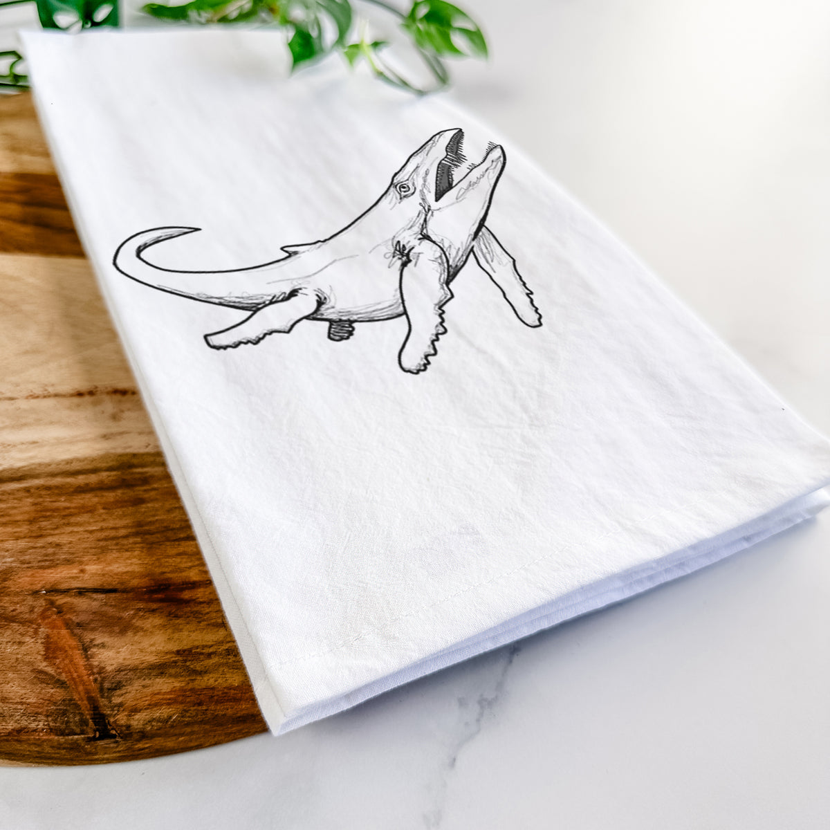 Close-up of a folded white tea towel on a wooden board, showing a black ink illustration of a Kronosaurus marine reptile swimming.
