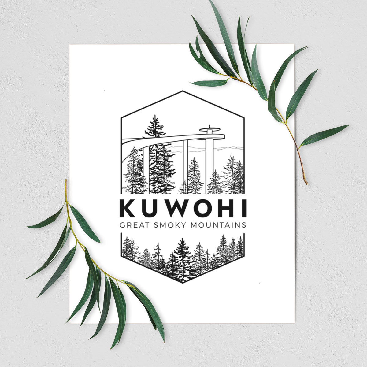 Art print of a black line art badge of the Kuwohi observation tower above pine trees, with the words Kuwohi, Great Smoky Mountains, shown with eucalyptus leaves.