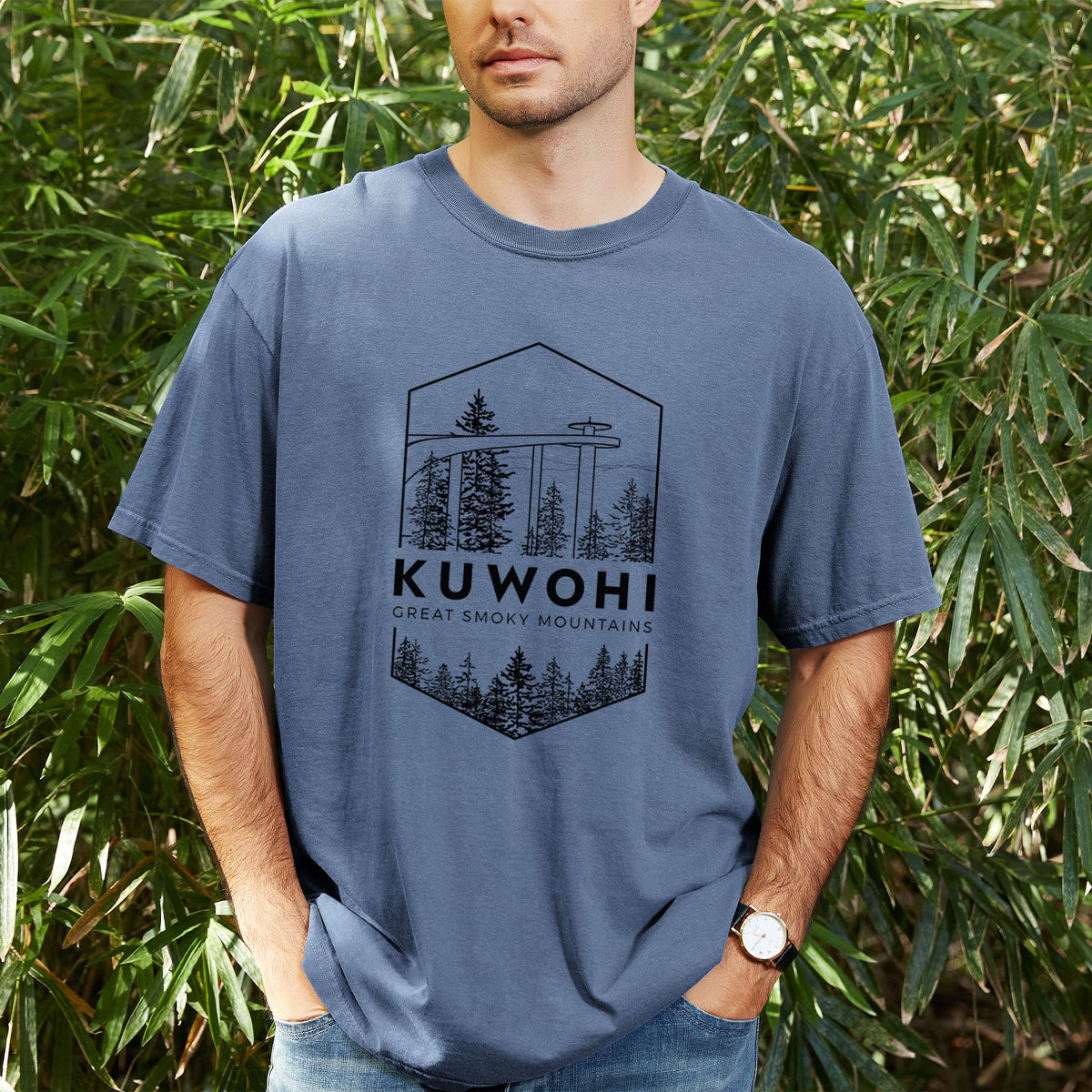 Man wearing a Comfort Colors t-shirt printed with a black line art badge of the Kuwohi observation tower above pine trees, with the words Kuwohi, Great Smoky Mountains, outdoors.