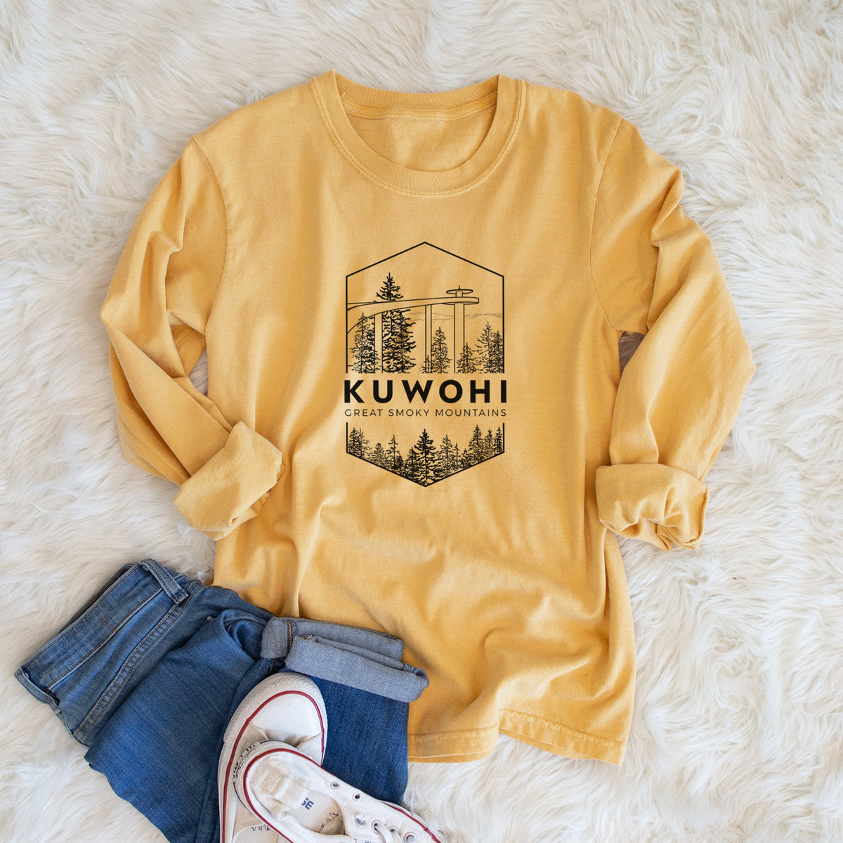 Golden wheat yellow Comfort Colors long sleeve shirt printed with a black line art badge of the Kuwohi observation tower above pine trees, with the words Kuwohi, Great Smoky Mountains, shown in a lifestyle photo.
