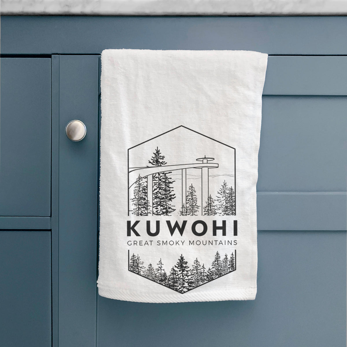 White hand towel printed with a black line art badge of the Kuwohi observation tower above pine trees, with the words Kuwohi, Great Smoky Mountains, hanging on a cabinet door.
