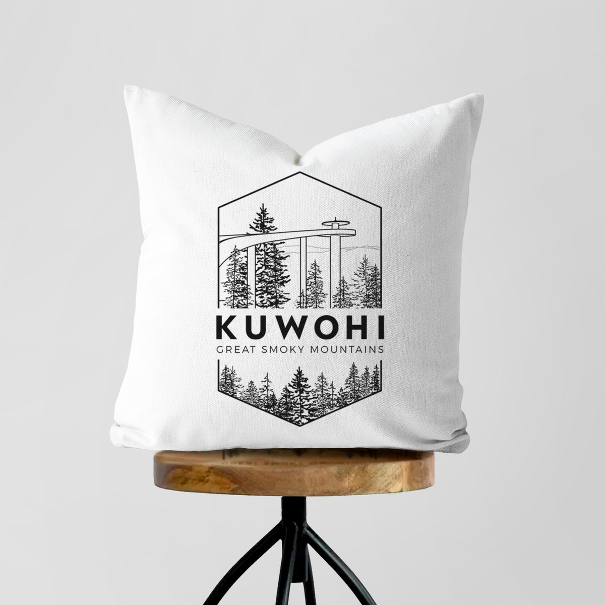 White pillow cover printed with a black line art badge of the Kuwohi observation tower above pine trees, with the words Kuwohi, Great Smoky Mountains, on a wooden stool.