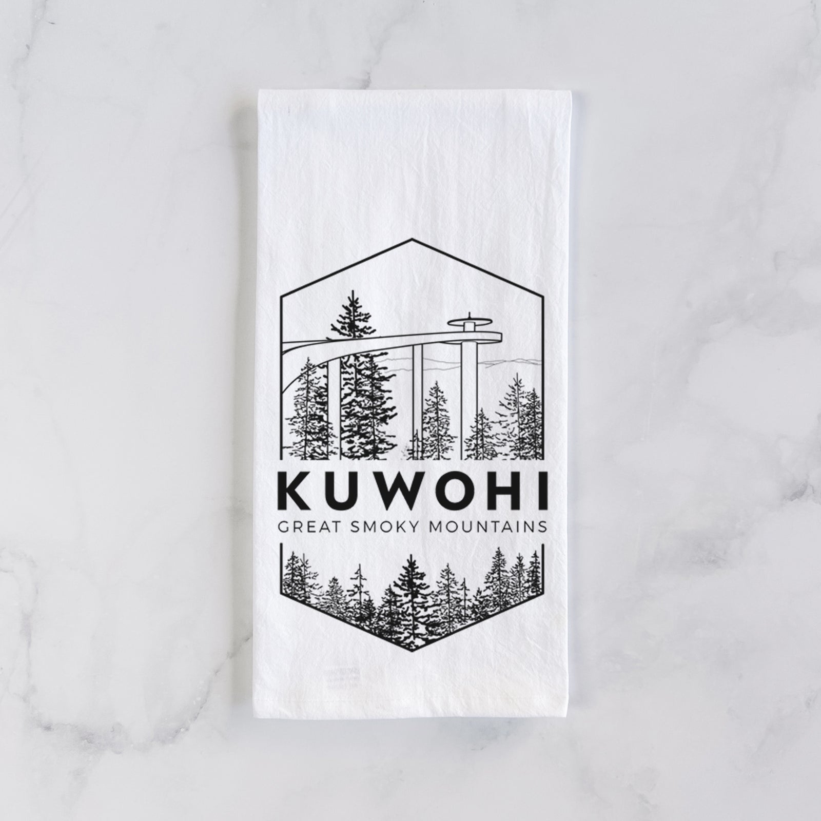 White cotton tea towel printed with a black line art badge of the Kuwohi observation tower above pine trees, with the words Kuwohi, Great Smoky Mountains, on a marble counter beside a round wooden cutting board and greenery.