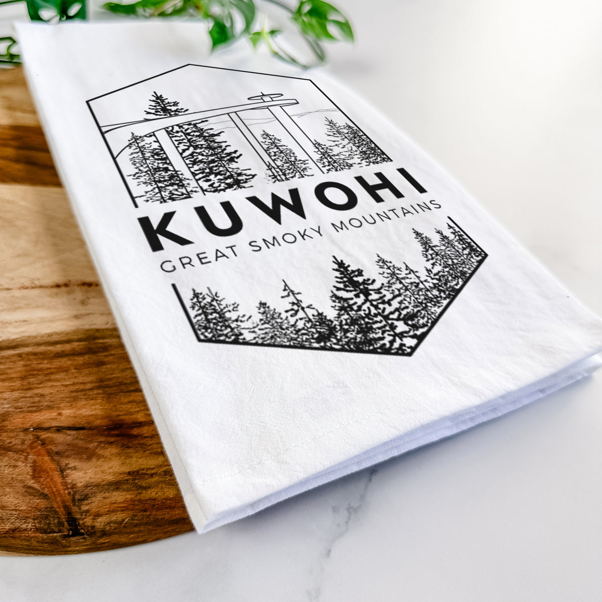 Close-up of a folded white tea towel on a wooden board, showing a black line art badge of the Kuwohi observation tower above pine trees, with the words Kuwohi, Great Smoky Mountains.